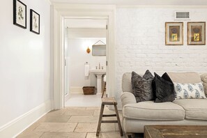 Una sala de estar con un sofá, una mesa de centro y un taburete de madera. Se aprecia una pared de ladrillo blanco con cuadros enmarcados y una puerta blanca que da a un baño.