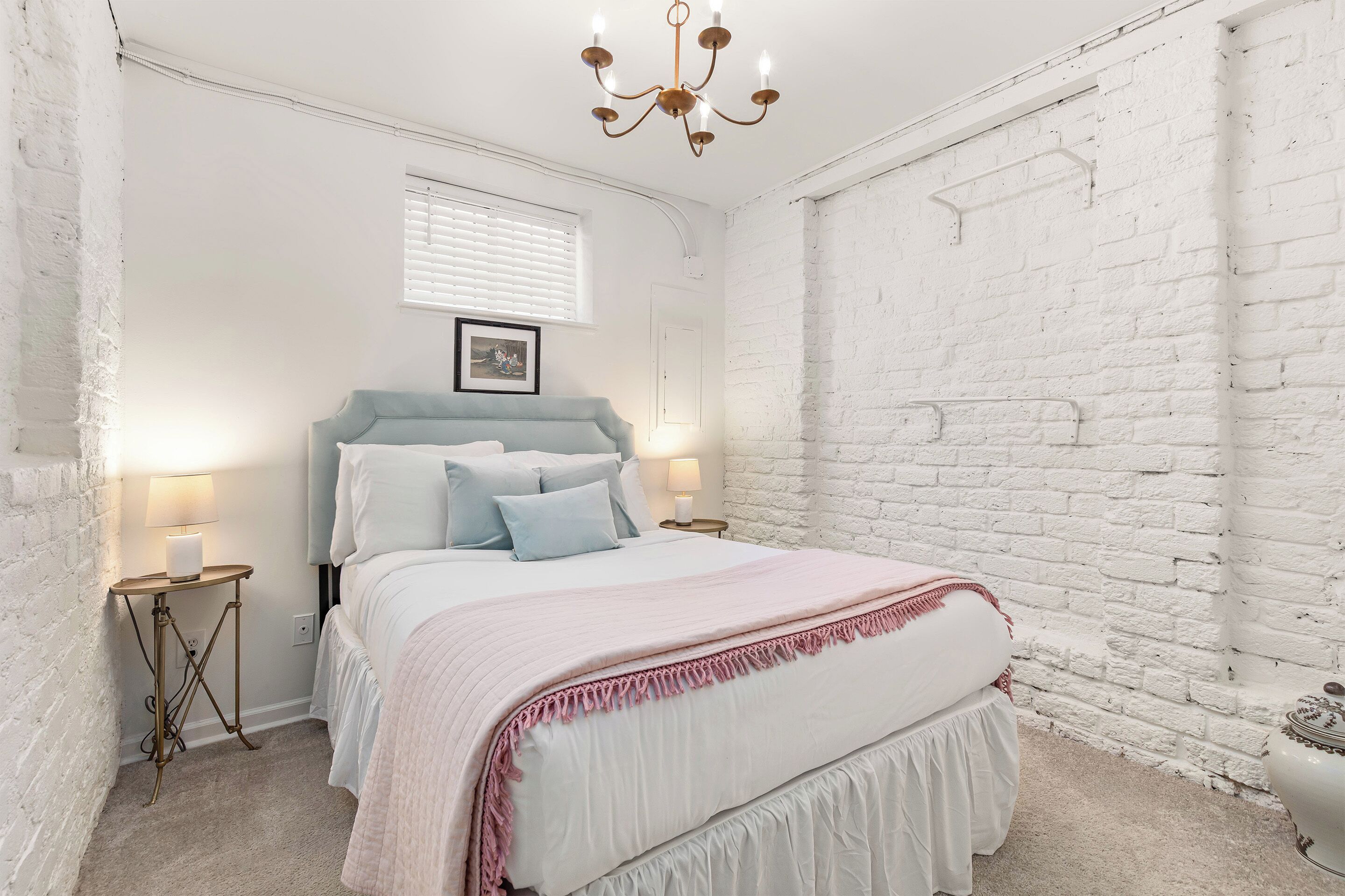 Een slaapkamer met een bed, nachtkastjes, een kroonluchter, een raam met zonwering en een witte baksteenmuur.