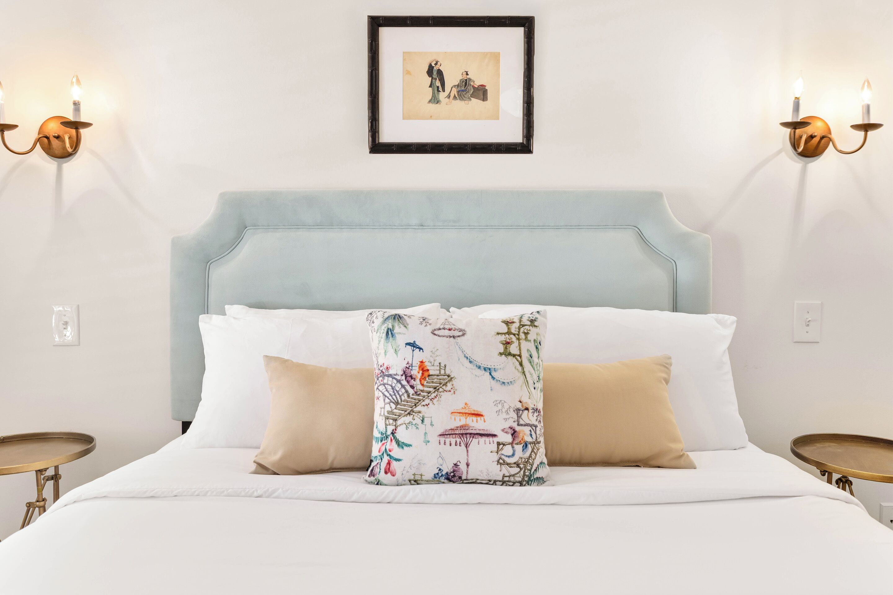 Een bed met een lichtblauwe hoofdbord, wit beddengoed en een decoratief kussen met een natuurscène. Twee wandlampen met kaarsen, en een ingelijst schilderij boven het bed.