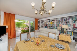 Ein helles Zimmer mit einem Esstisch für zwei Personen, einem weißen Sofa und einem großen Bücherregal, gefüllt mit Büchern.