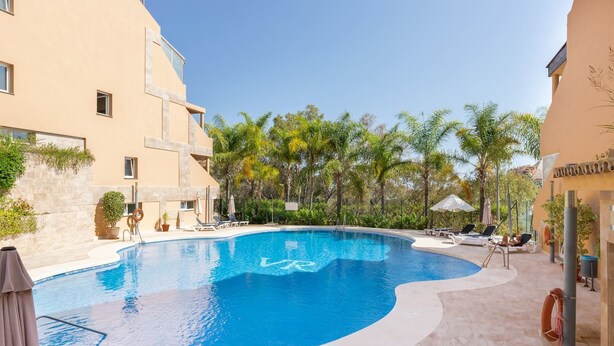 A swimming pool with lounge chairs and umbrellas, surrounded by palm trees and a building with a beige facade. - Real1b - Beautiful apt With Tropical Terrace (Marbella)
