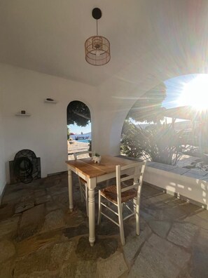 Ein Zimmer mit Steinfußboden, einem Holztisch, weißen Stühlen, einer Hängeleuchte und Blick ins Freie.