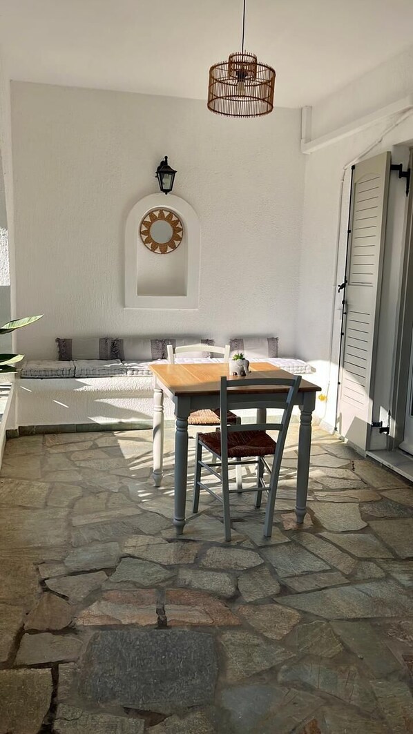 Eine Terrasse mit Steinboden, einem Holztisch und -stühlen, einer hängenden Leuchte und einem wandmontierten Ziertegels.