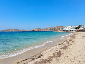 A beach with clear blue waters, sandy shore, and white buildings in the background. - Paros Casa Prive - Poseidon Shared Pool Nest (Ag Charalambos)