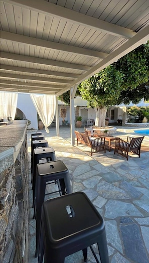 A covered outdoor seating area with stone flooring, a pool, and a tree providing shade. - Paros Casa Prive - Poseidon Shared Pool Nest (Ag Charalambos)