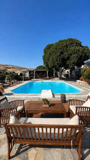 A pool with lounge chairs and a table, surrounded by stone flooring and a large tree. - Paros Casa Prive - Poseidon Shared Pool Nest (Ag Charalambos)