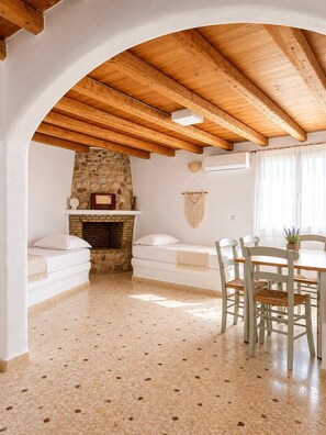 A room with a stone fireplace, wooden ceiling beams, a dining table with chairs, and a white sofa. - Paros Casa Prive - Deluxe Shared Pool Nest (Ag Charalambos)
