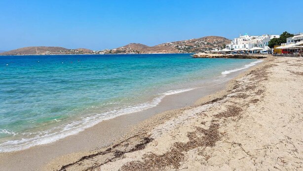 Una playa con aguas azules cristalinas, orilla de arena y edificios blancos al fondo.
