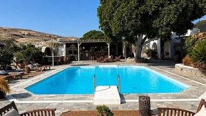 Piscina con sillas de descanso y una mesa, rodeada de suelo de piedra y un árbol grande.