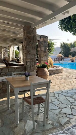 A stone patio with a table and chairs, a pool, and a stone wall. - Paros Casa Prive - Iria Shared Pool Nest (Ag. Charalampos)