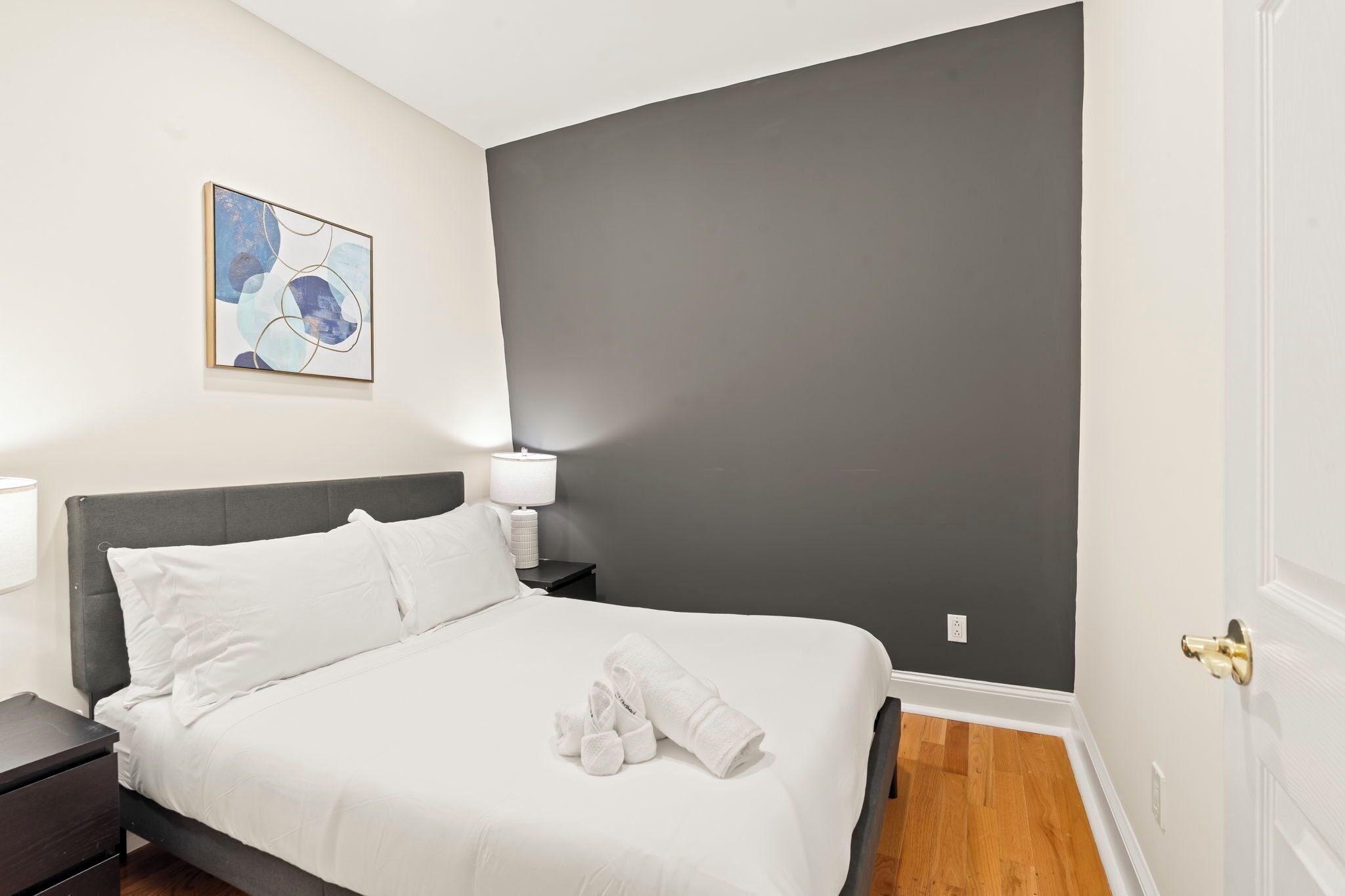 A neatly made bed with white linens and a rolled towel, a bedside table with a lamp, and a wall-mounted abstract painting.