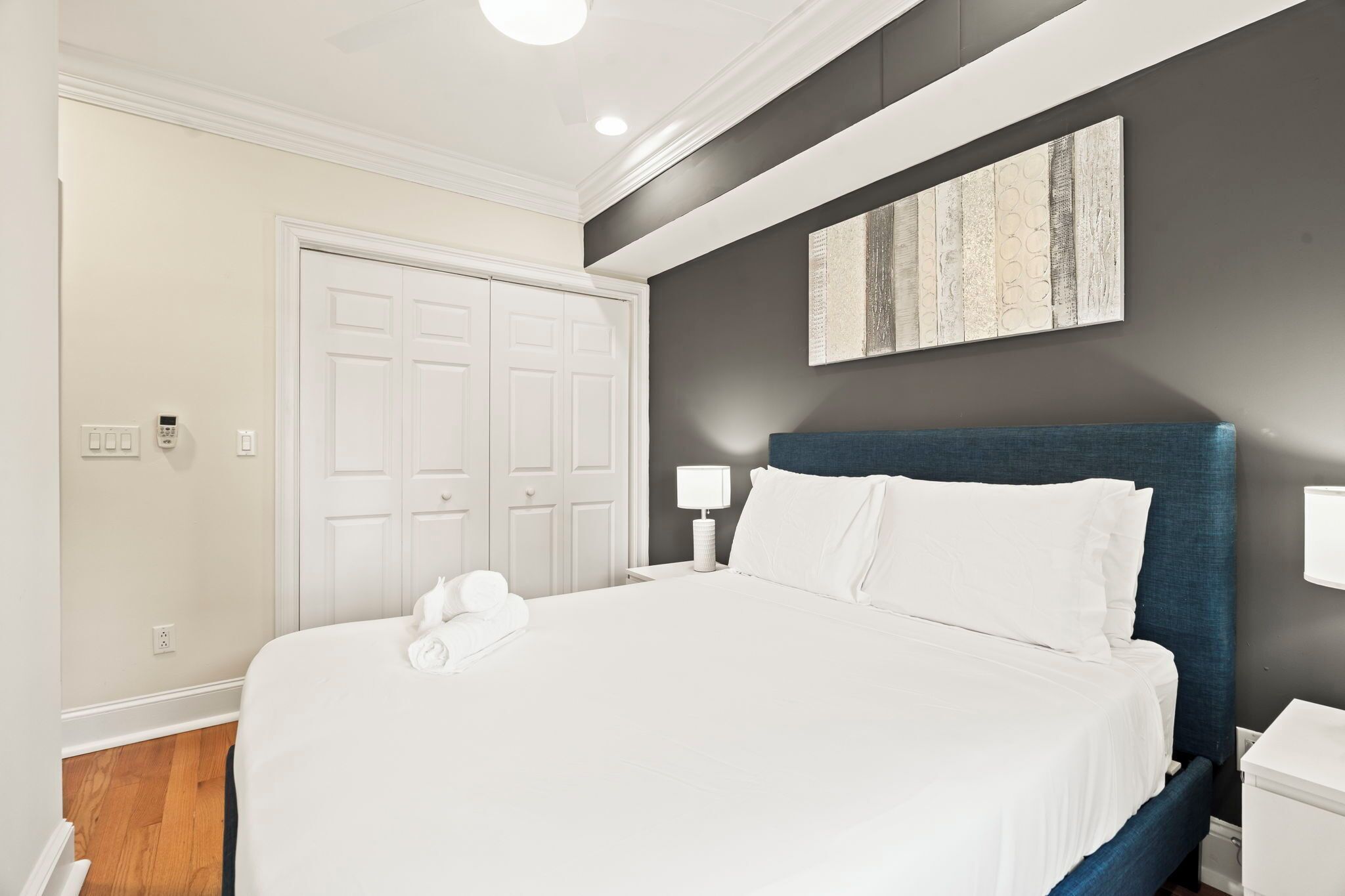 A neatly made bed with white linens, a blue headboard, a white bedside table, and a wall-mounted artwork.