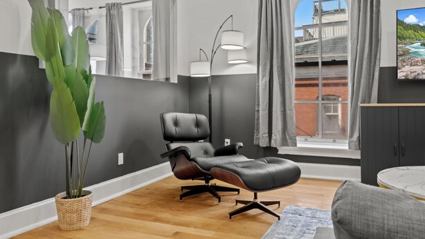 Een moderne woonkamer met een leren fauteuil, een grote potplant, een vloerlamp en uitzicht op een brug door het raam.