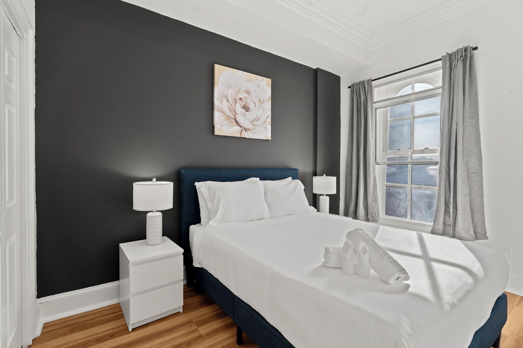 A bedroom with a dark wall, a white bedside table, a blue headboard, a white bed, a window with curtains, and a painting on the wall.