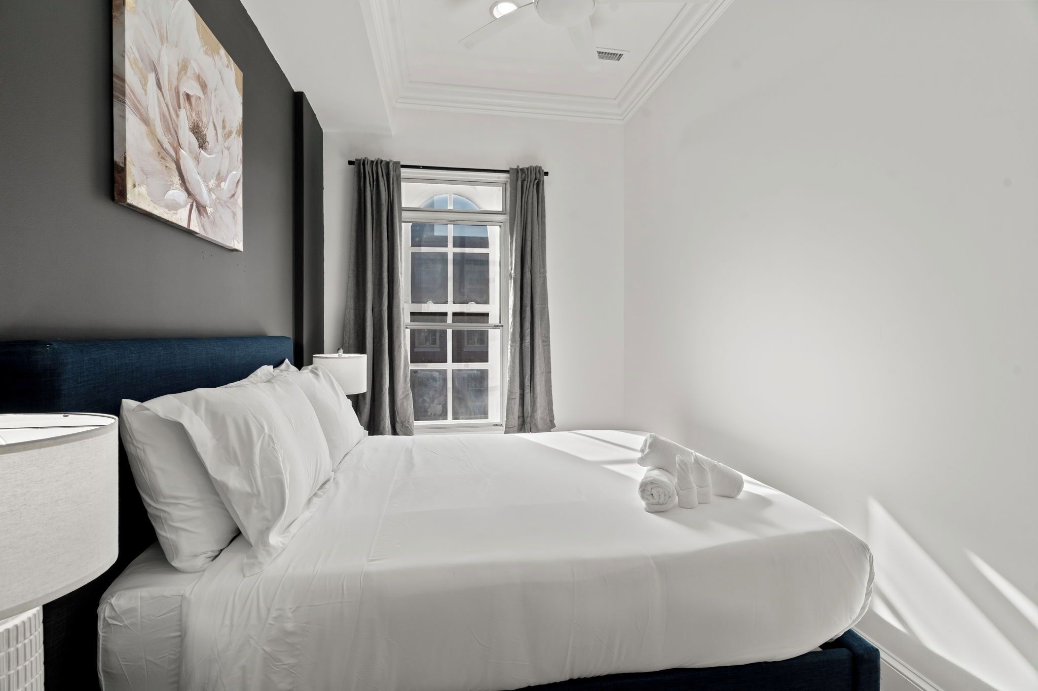 Una camera d'albergo con un letto grande, una finestra con tende, una ventola a soffitto e un'opera d'arte appesa al muro.