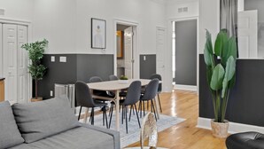 Een moderne woonkamer met een grijze bank, een eettafel met stoelen, een grote potplant en een zwarte lederen fauteuil.
