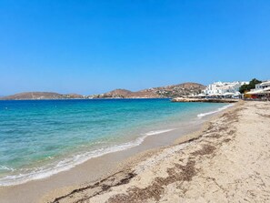 Una playa con aguas azules cristalinas, orilla de arena y edificios blancos al fondo.