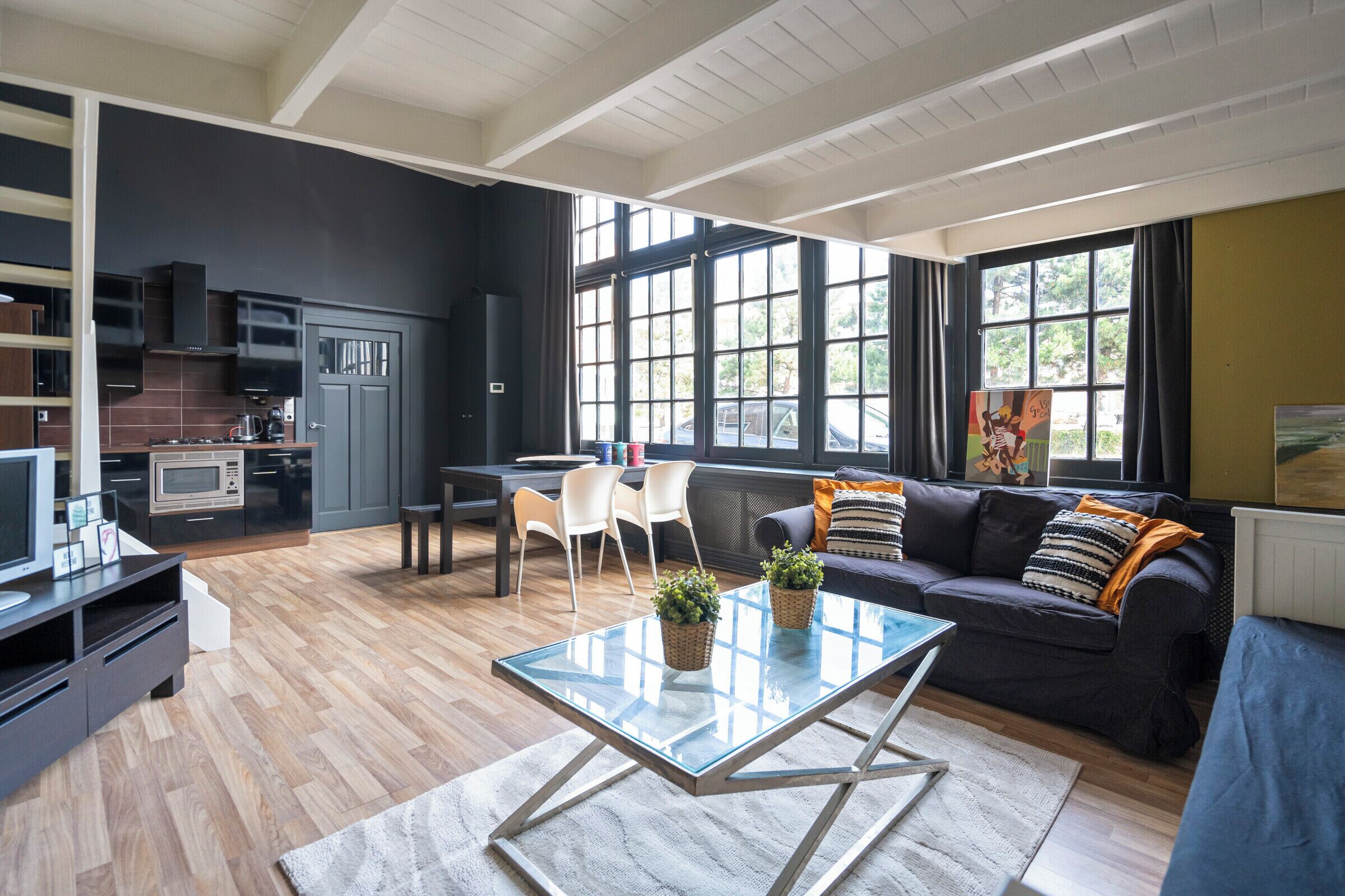 A modern living room with a glass coffee table, a dark-colored sofa, and a kitchen area with a microwave and stove.
