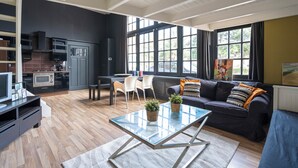 Ein modernes Wohnzimmer mit einem Glastisch, einem dunkelgrauen Sofa und einer Küchenzeile mit Mikrowelle und Herd.