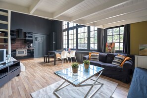 A modern living room with a glass coffee table, a dark-colored sofa, and a kitchen area with a microwave and stove. - De IJsfabriek - Industrial loft studio in Zandvoort (Zandvoort)
