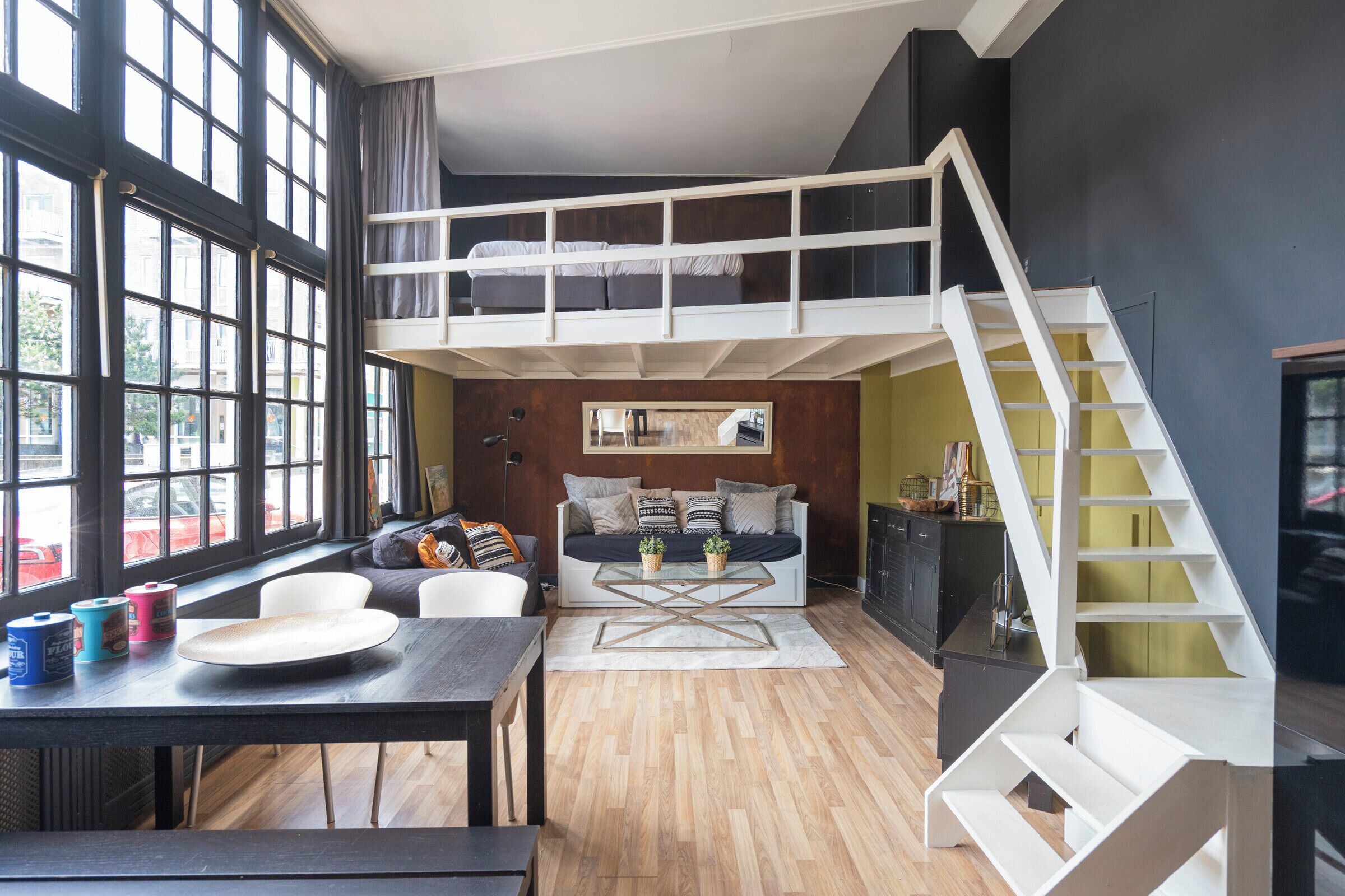 Un espacio tipo loft con techo alto, una escalera, un sofá, una mesa de comedor y un ventanal grande.