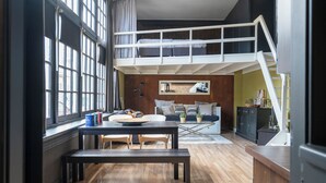 Ein Loft-Zimmer mit Stockbett, Esstisch mit Stühlen, Küchenzeile und großem Fenster.