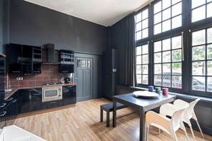 A modern kitchen with dark cabinetry, a built-in microwave, and a dining area with a table and chairs. - De IJsfabriek - Industrial loft studio in Zandvoort (Zandvoort)