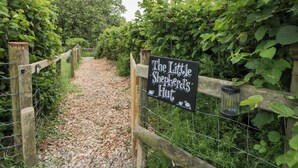 Ein Kiesweg führt zu einem Schild mit der Aufschrift 'The Little Shepherd's Hut', begleitet von einer Holzlattenzaun und üppiger Bepflanzung an beiden Seiten.
