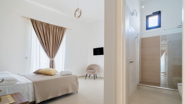 Quarto com cama, cadeira, mesa de cabeceira, janela com cortinas e área de chuveiro com porta de vidro.