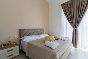 Una cama bien hecha con cabecera, una mesita de noche con lámpara y una ventana con cortinas.