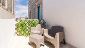 Varanda com duas cadeiras e uma mesa pequena, grade com vegetação e um prédio com porta azul.