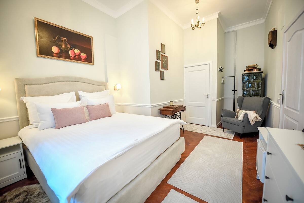 A bedroom with a large bed, a nightstand, a dresser, a chair, a small table, a chandelier, and a painting on the wall.
