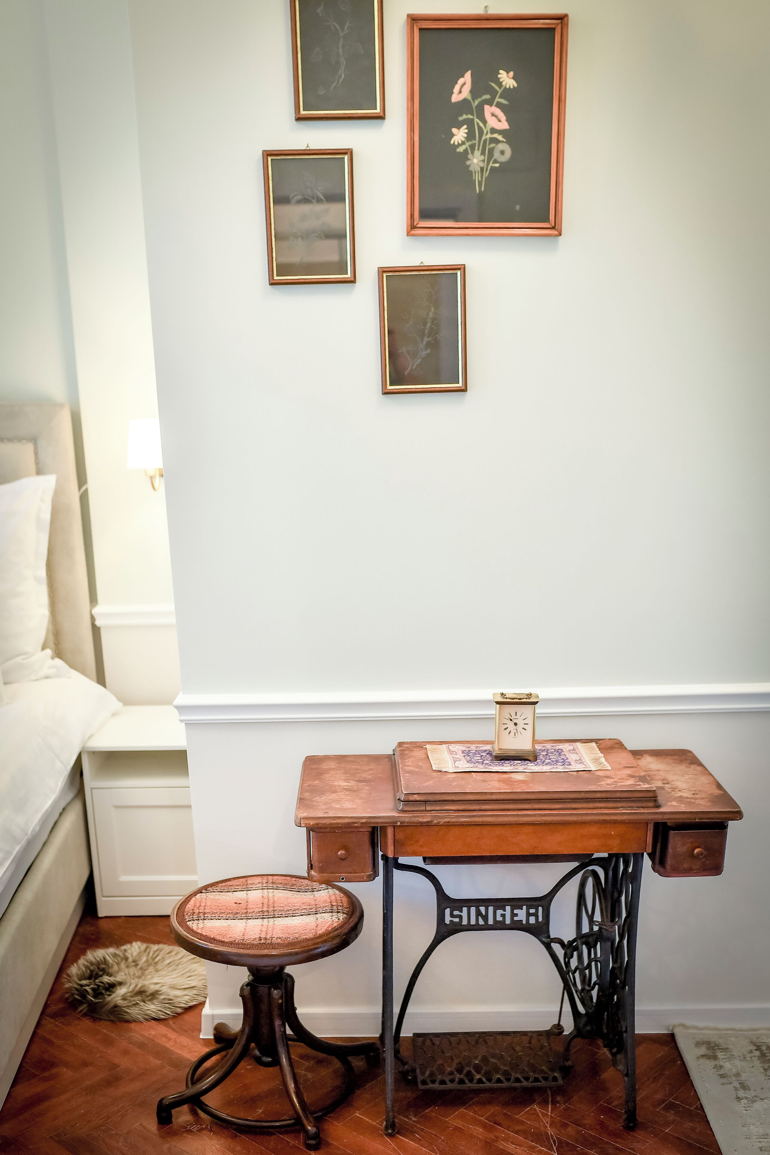 A room with a bed, a desk with a sewing machine, a small stool, and framed pictures on the wall.