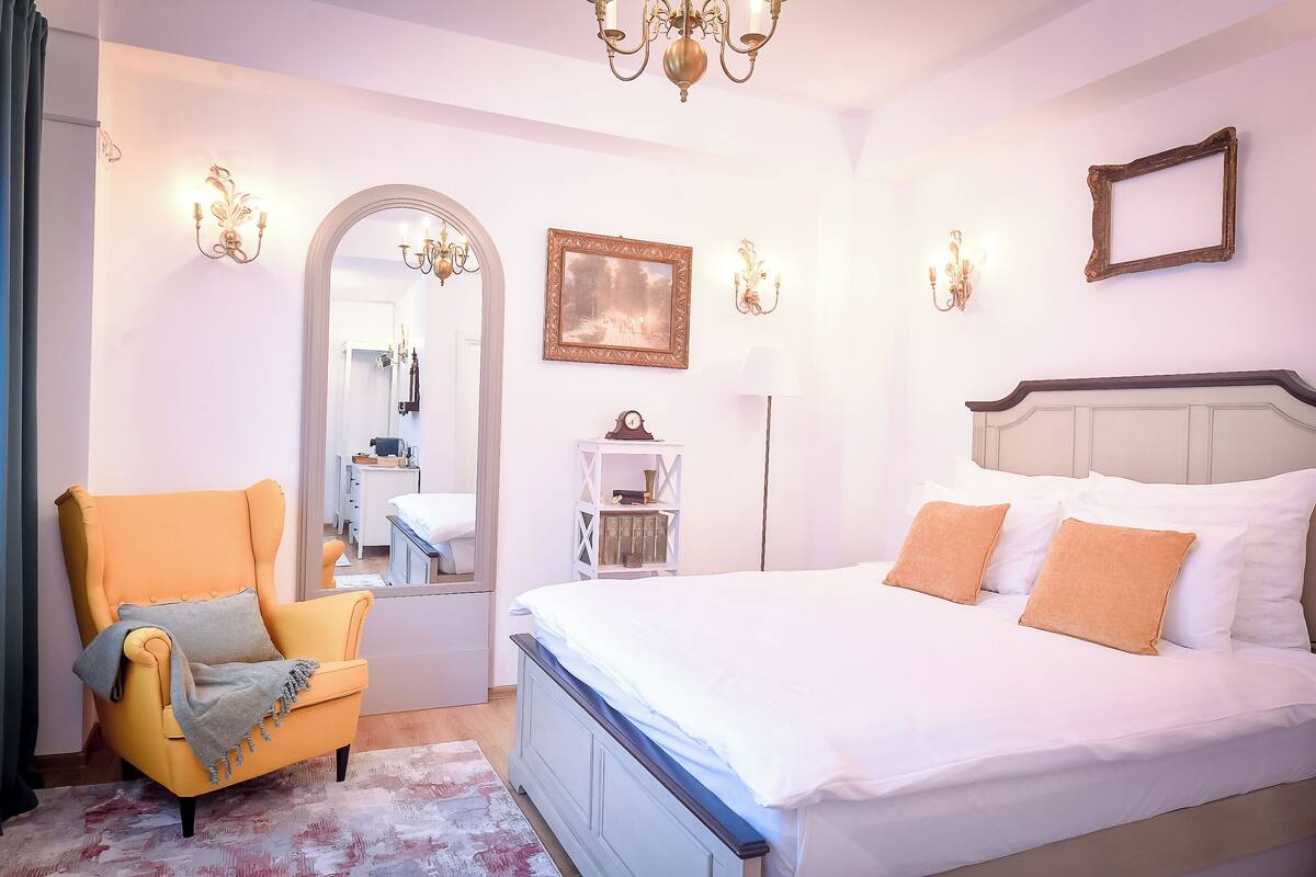 A bedroom with a bed, a chair, a mirror, and wall sconces.