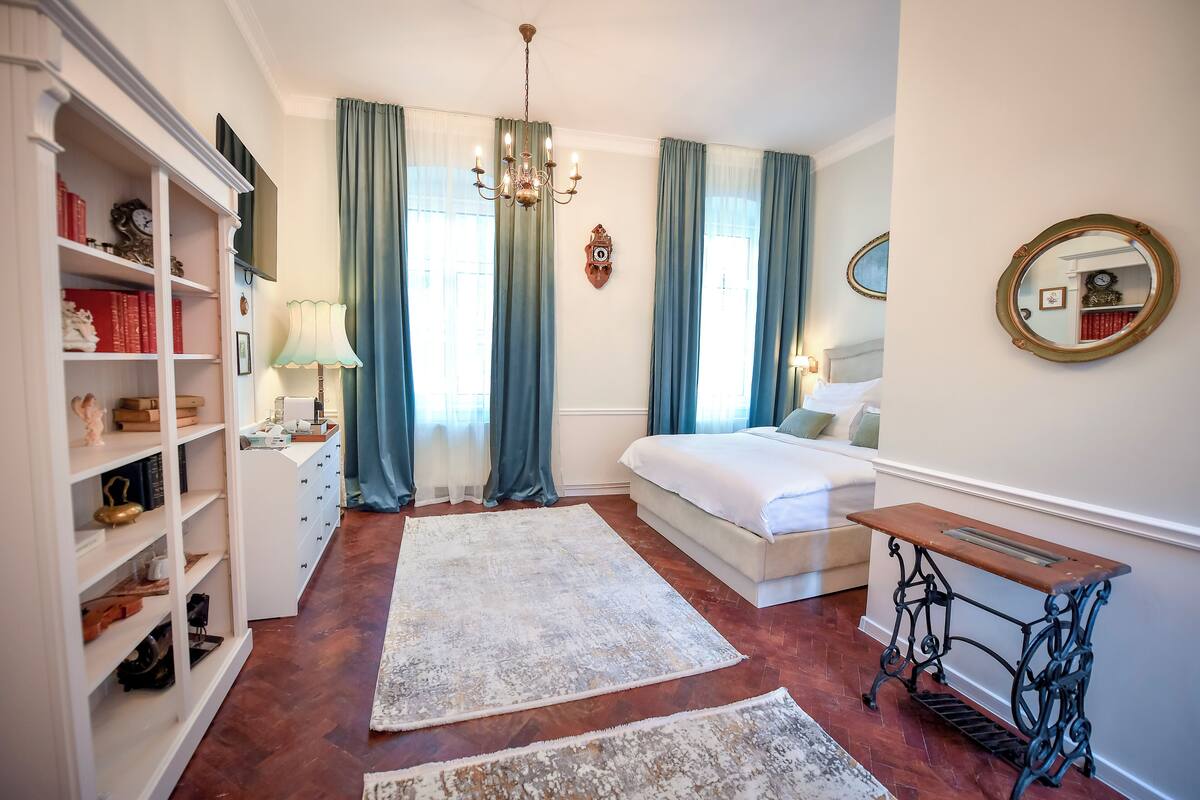 A bedroom with a bed, a sewing machine, a bookshelf, and a large window with curtains.
