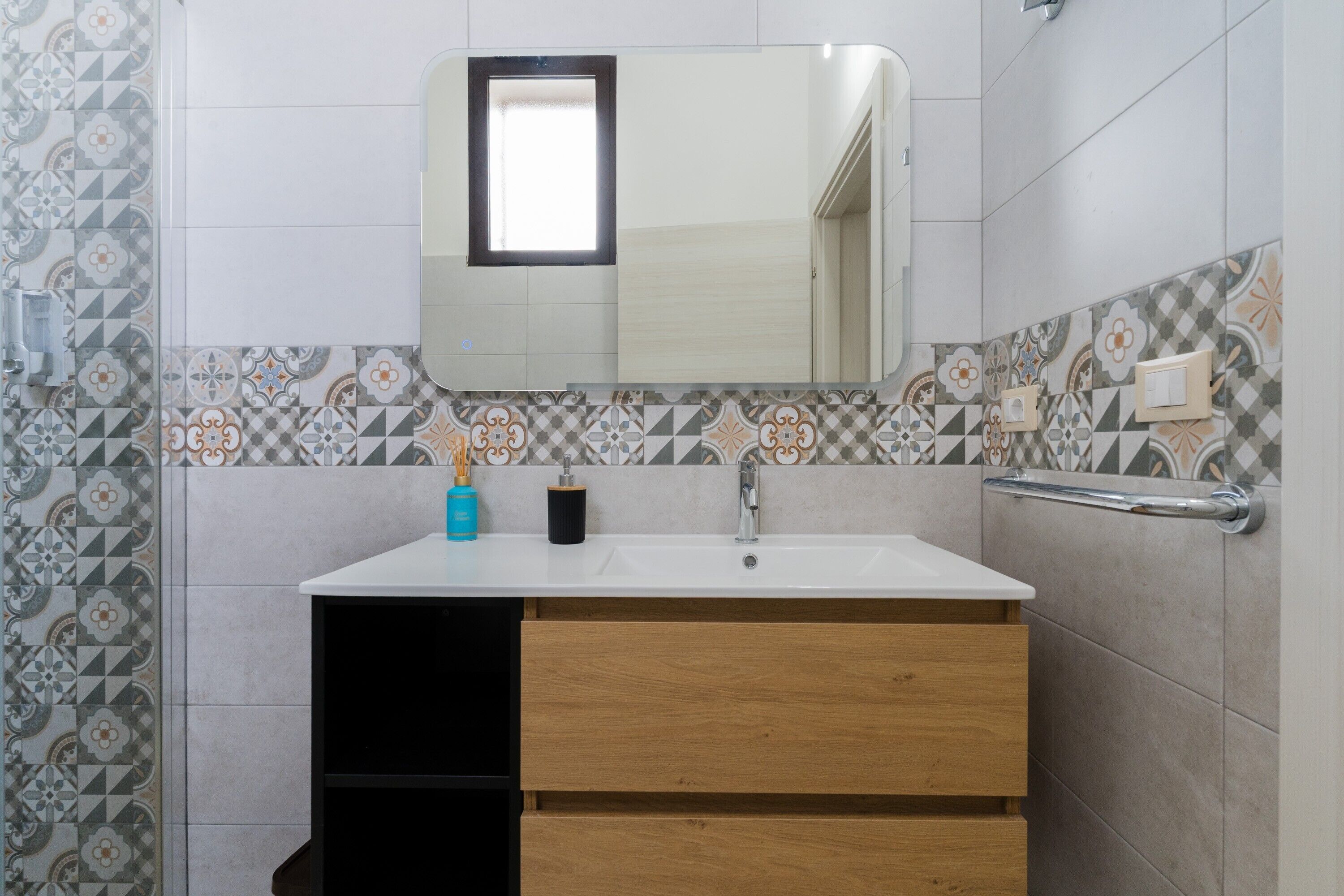 Une salle de bain avec un lavabo blanc, des tiroirs en bois, un miroir agrémenté d’une petite fenêtre et des carreaux à motifs.