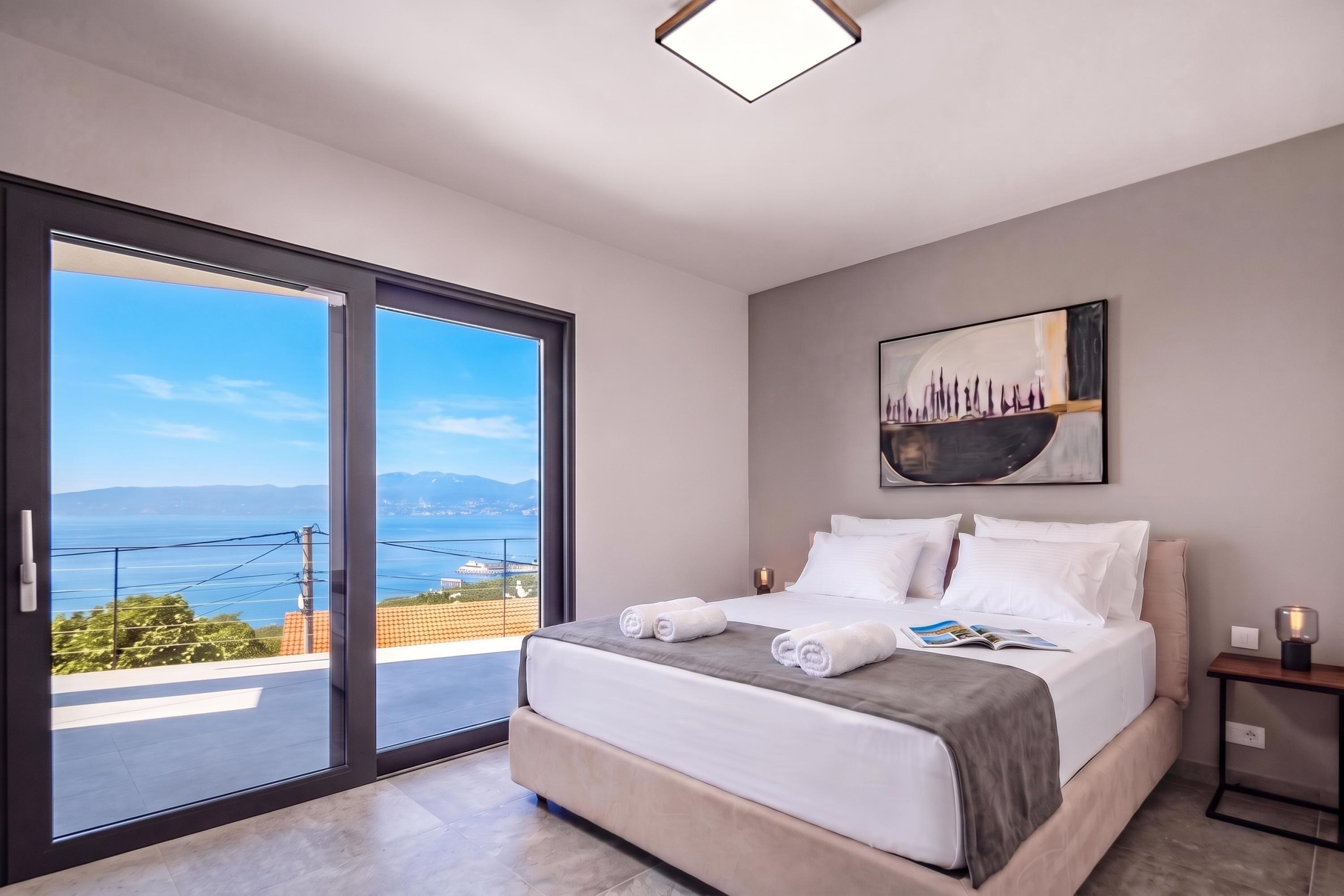 A bedroom with a large sliding glass door leading to a balcony, a bed with white linens, a painting on the wall, and a nightstand with a lamp.