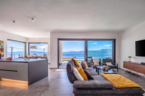 A modern living room with a large sofa, a coffee table, and a TV. There is a kitchen area with a bar and a view of the sea. - Pool Villa Solea /w Panoramic Sea View (Kostrena)