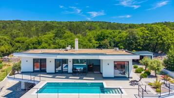 A modern two-story house with a swimming pool, large windows, and a view of a forested hill.
