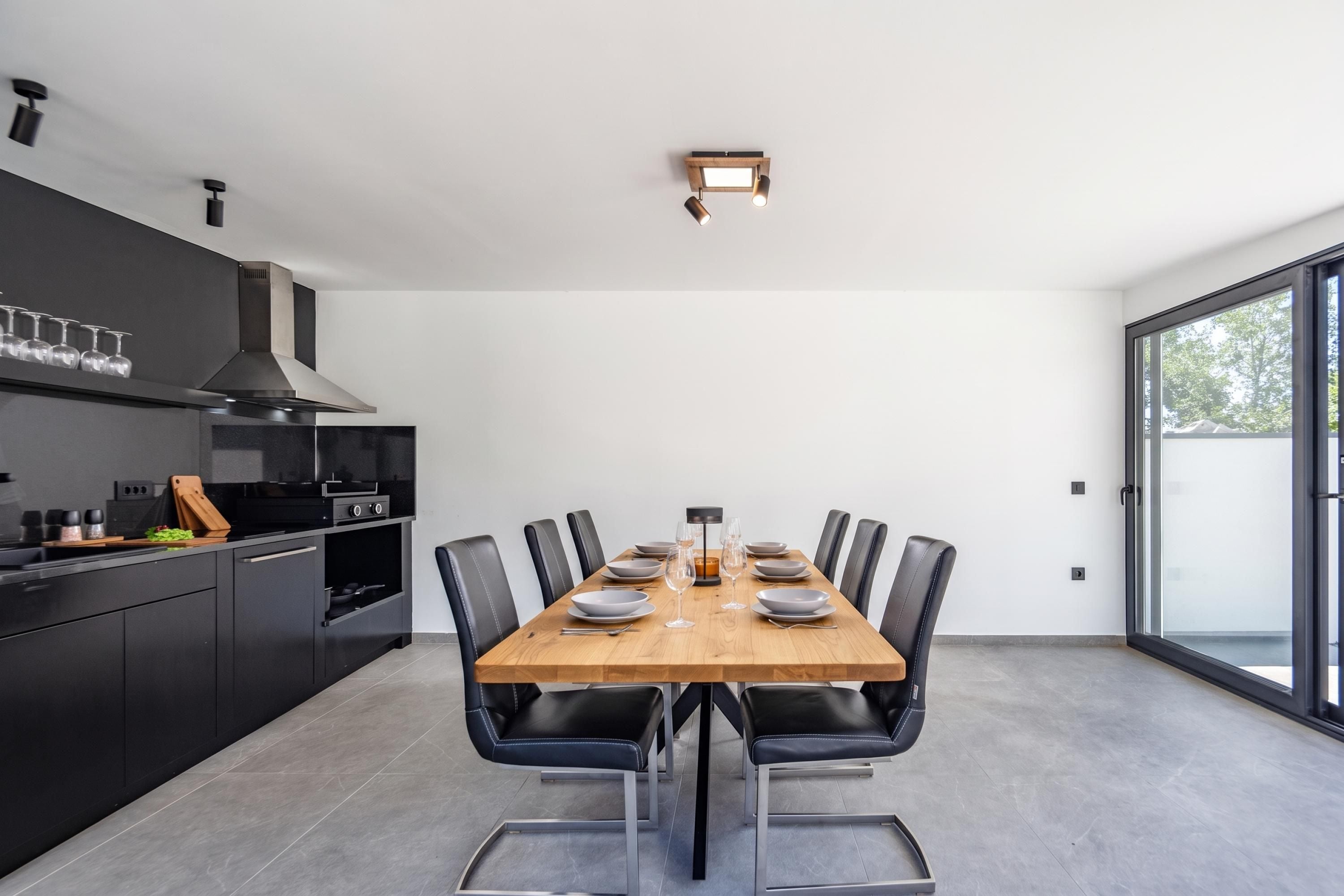 A modern kitchen with a dining area, featuring a large table, black chairs, and a view through a glass door.