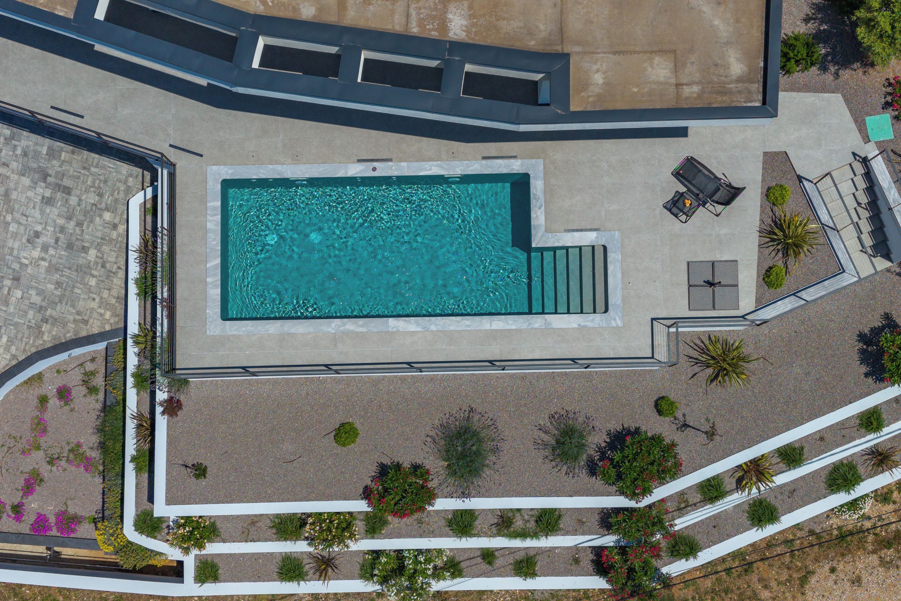 An aerial view of a swimming pool with a patio area, surrounded by landscaped gardens and a stone wall.