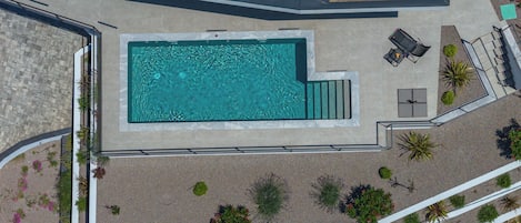 An aerial view of a swimming pool with a patio area, surrounded by landscaped gardens and a stone wall.