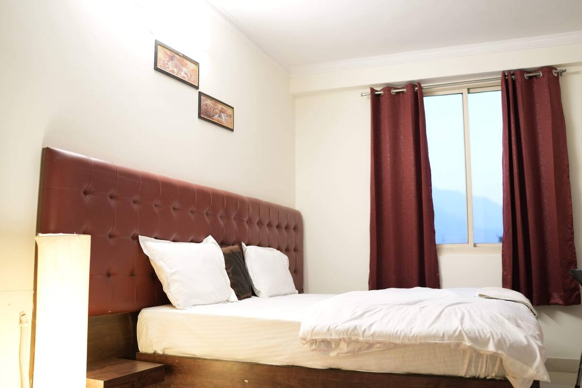 Un dormitorio con una cama grande, cabecera, una ventana con cortinas y una mesita de noche.