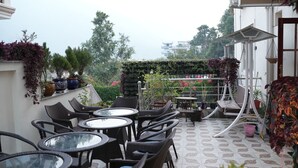 An outdoor seating area with wicker chairs and tables, surrounded by potted plants and a patterned floor. - Ayuskama Ayurveda Ayur Yoga (Rishikesh)