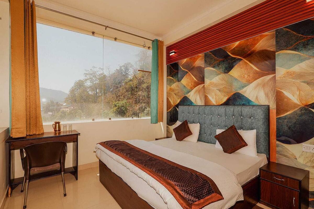 Habitación de hotel con cama, escritorio, silla, mesita de noche y un gran ventanal con vista a árboles.
