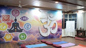 A yoga studio with mats, cushions, a wall with yoga symbols, a ceiling fan, and a window. - Ayuskama Ayurveda Ayur Yoga (Rishikesh)