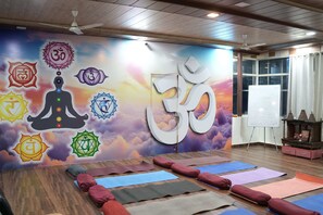 A yoga studio with mats, cushions, a wall with yoga symbols, a ceiling fan, and a window. - Ayuskama Ayurveda Ayur Yoga (Rishikesh)