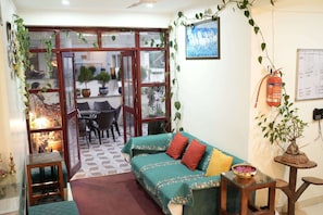 A cozy living room with a sofa, a small table, a potted plant, and a fire extinguisher on the wall. - Ayuskama Ayurveda Ayur Yoga (Rishikesh)