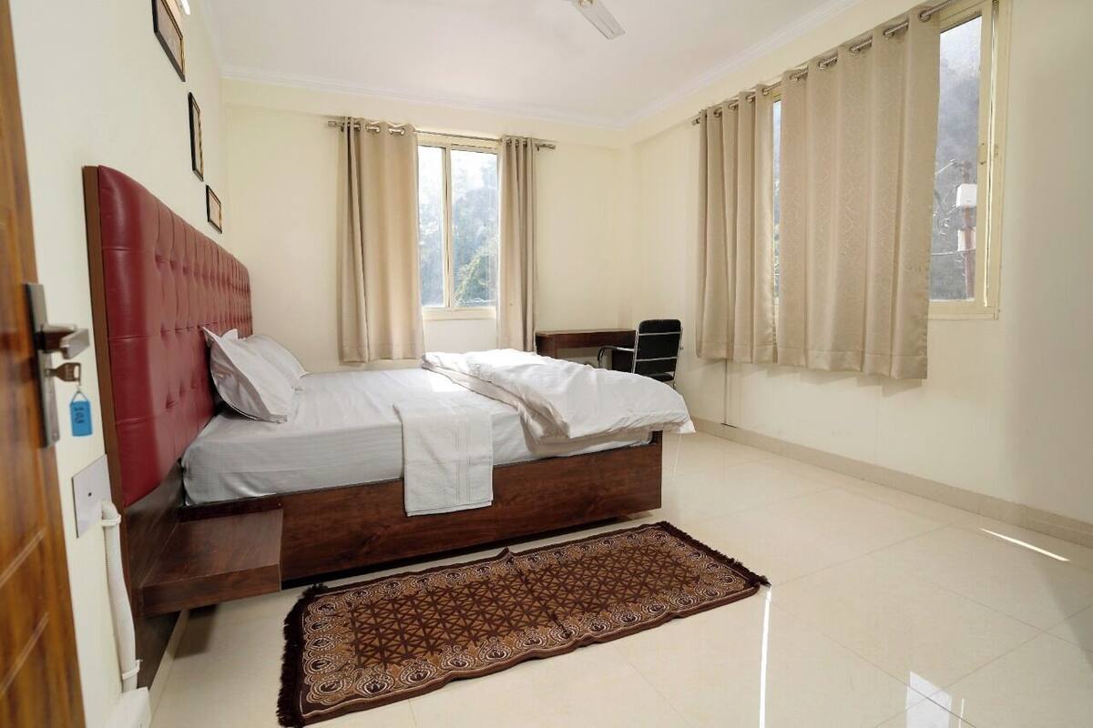 Un dormitorio con cama, cabecera de madera, ventana con cortinas, radiador y alfombra estampada.