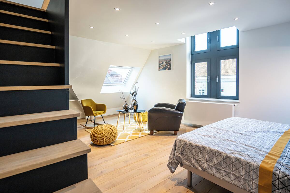 A modern bedroom with a bed, a chair, a small table, and a staircase.